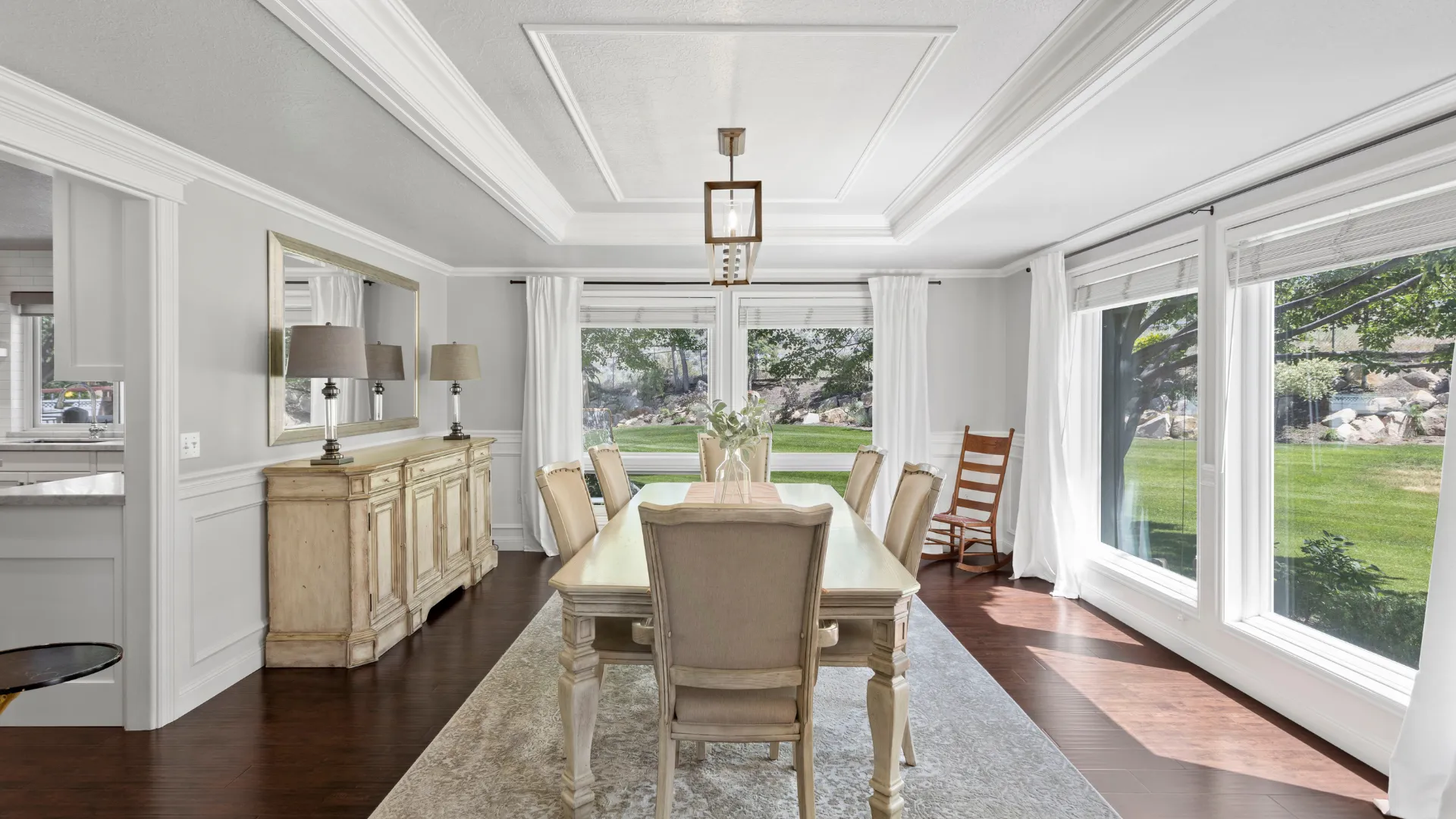 Dining room with beige table, chairs, rug, and big windows to garden.