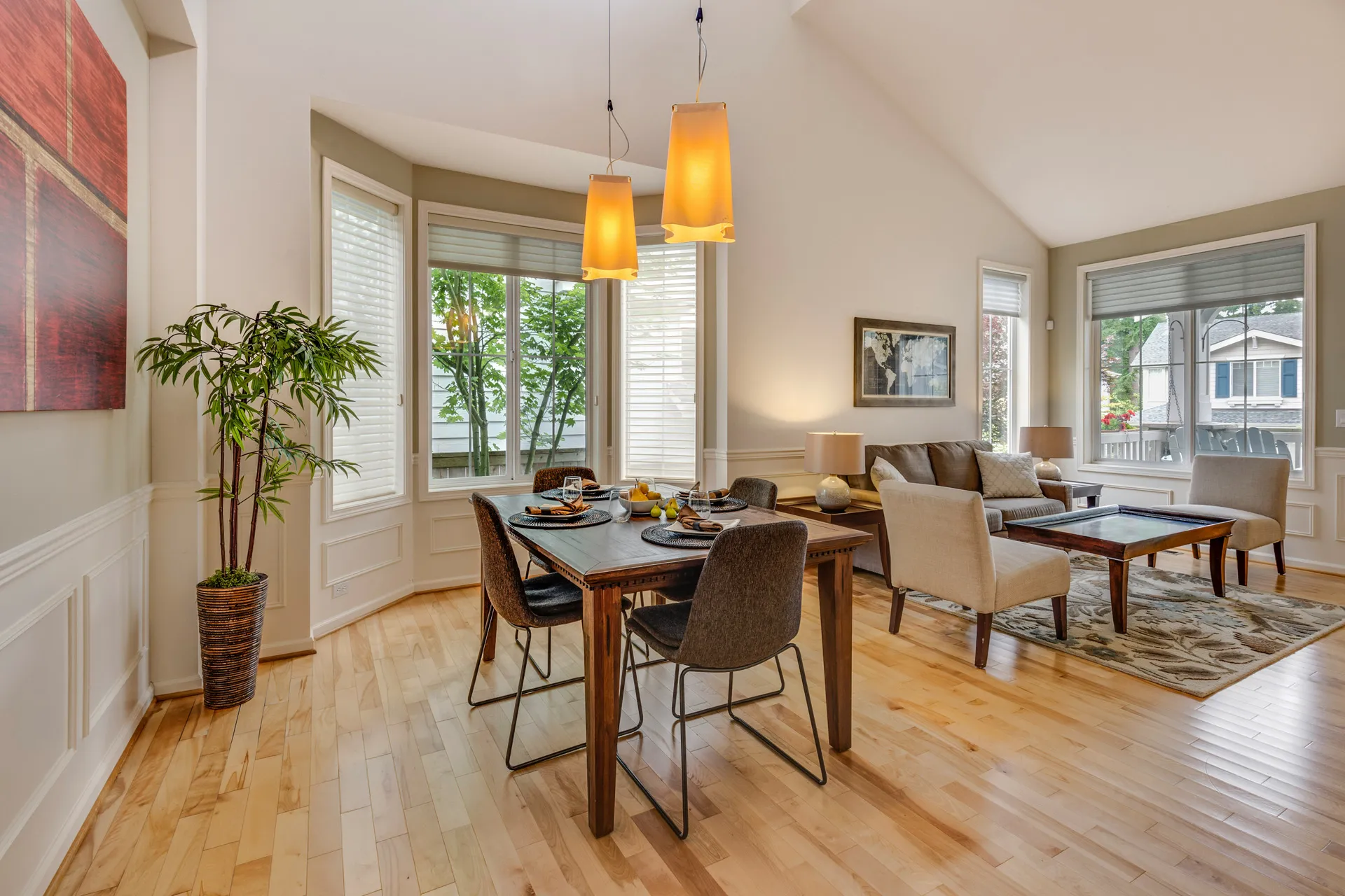 Dining area with wooden table, chairs, pendant lights, windows, sofa, and plants.