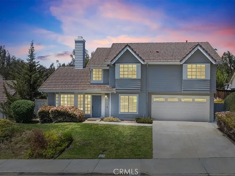 Two story house California real estate with blue walls, lit windows, bushes, and lawn.