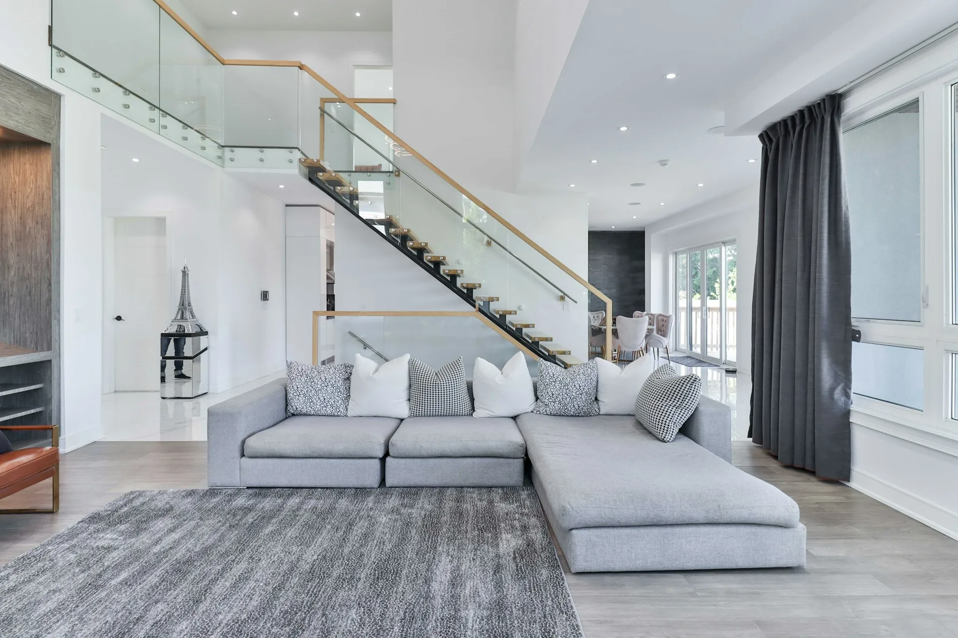 Living room with large gray sofa, rug, glass railing staircase, and floor-to-ceiling windows.